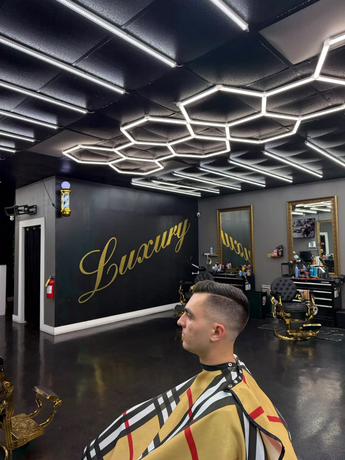 A client mid-cut beneath the hexagon lighting, the gold Luxury script painted on the wall behind him